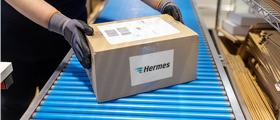 Person mit Handschuhen nimmt Paket mit Hermes-Versandetikett von einem Förderband. 
